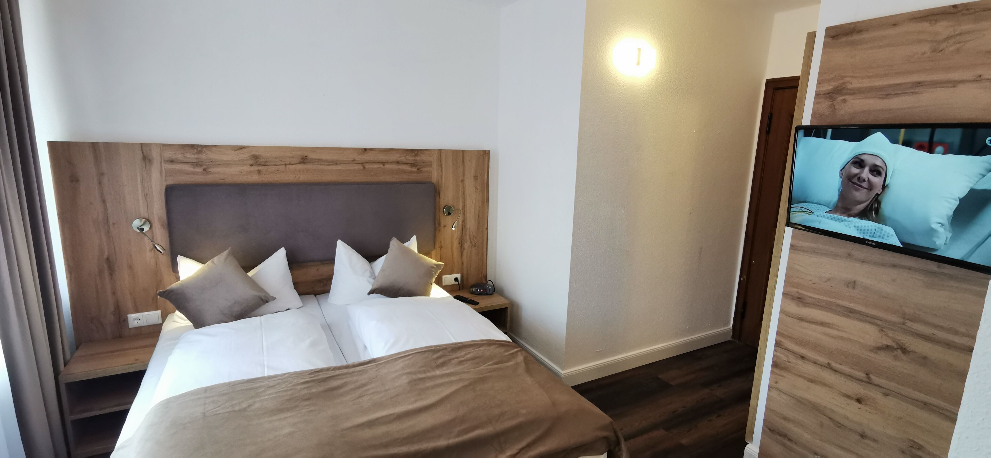A hotel room with a wooden headboard, a bed with white bedding and multiple pillows, a bedside table with a lamp, and a television mounted on the wall.