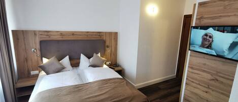 A hotel room with a wooden headboard, a bed with white bedding and multiple pillows, a bedside table with a lamp, and a television mounted on the wall.