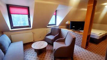 Ein kleiner, gemütlicher Raum mit einer Couch, zwei Sesseln, einem Couchtisch und einem Fernseher auf einem Schrank. Zwei Fenster mit Jalousien, ein Heizkörper und ein gemusterter Teppich sind ebenfalls vorhanden.