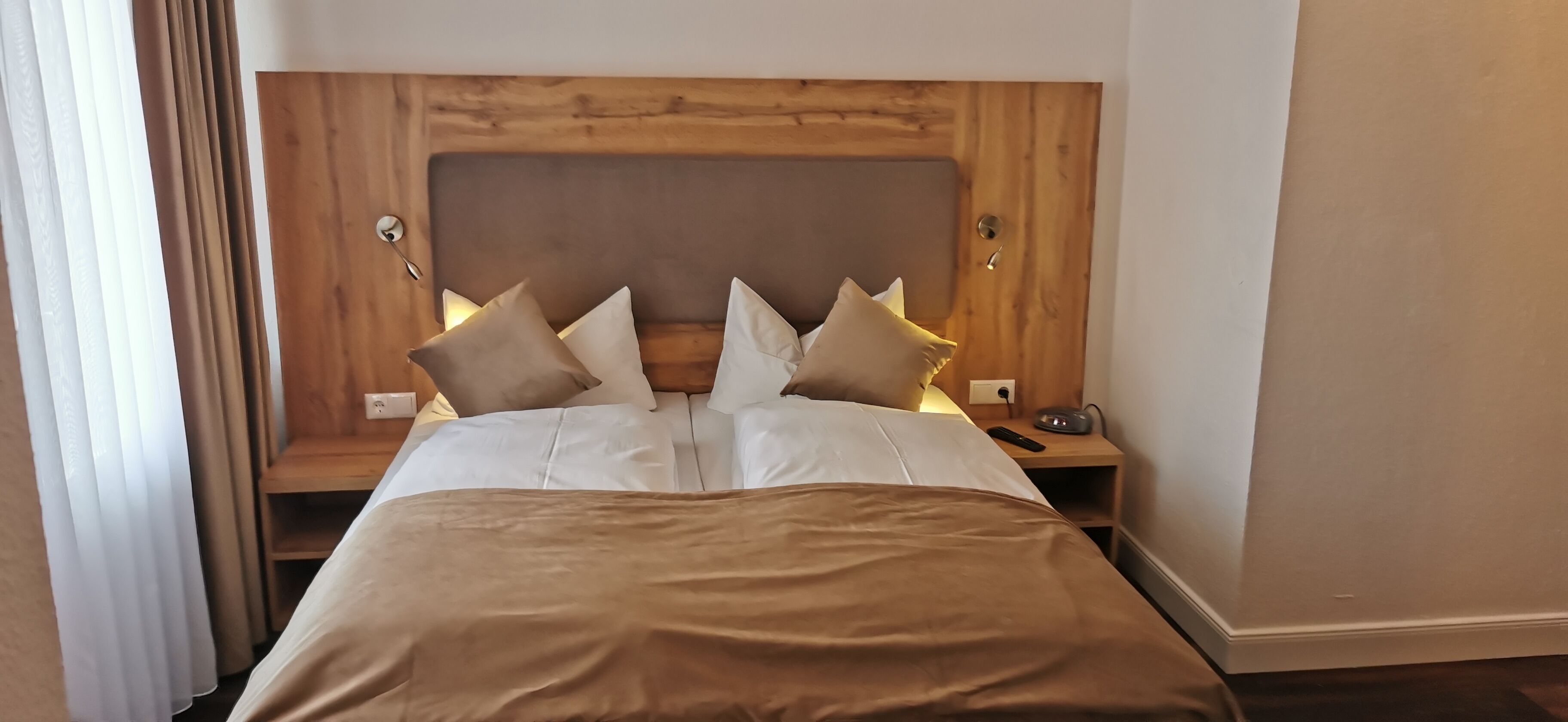 Una cama bien hecha con cabecera de madera, ropa de cama blanca y varias almohadas. Se aprecia una mesita de noche con una lámpara y un control remoto.