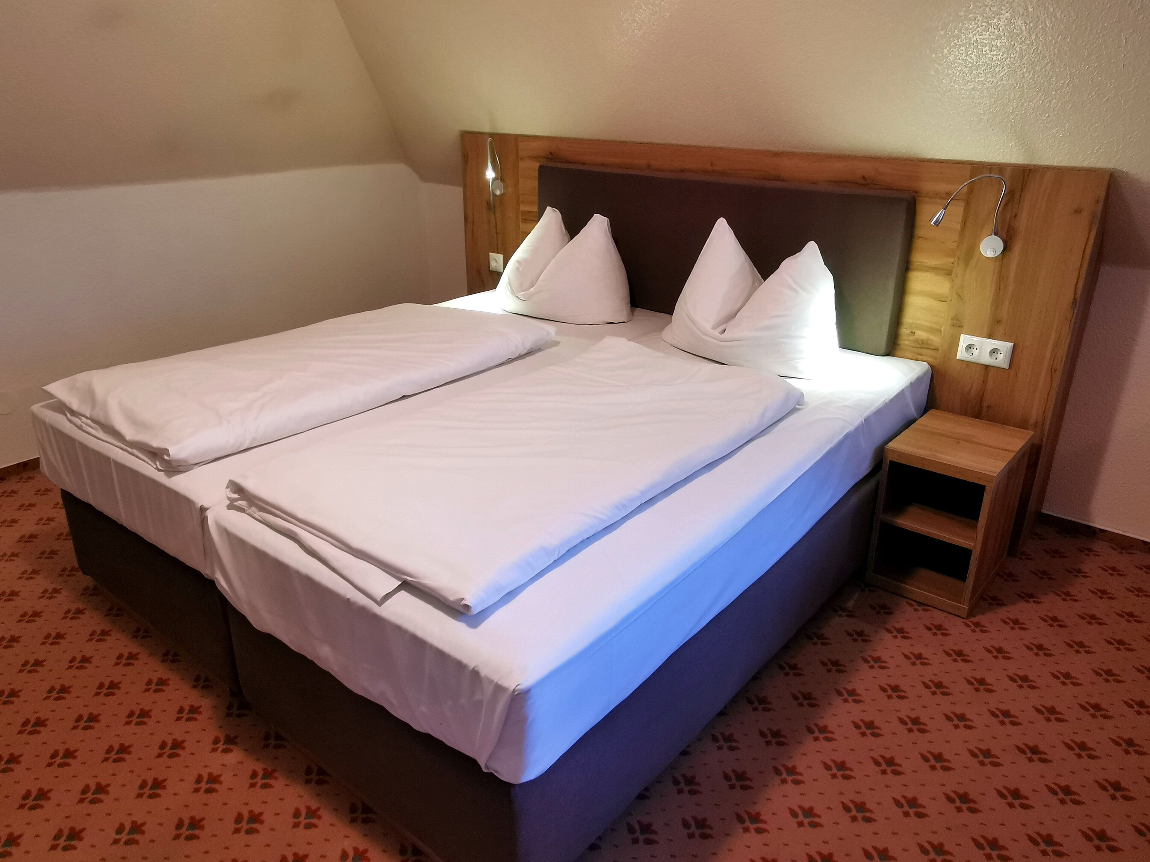 Un lit double avec du linge de lit blanc et des oreillers, une tête de lit en bois, une table de chevet et une moquette à motifs.