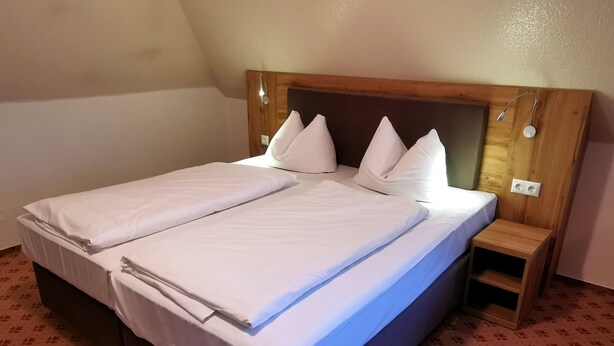 A double bed with white linens and pillows, a wooden headboard, a bedside table, and a patterned carpet. - Heidehotel Soltauer Hof (Soltau)