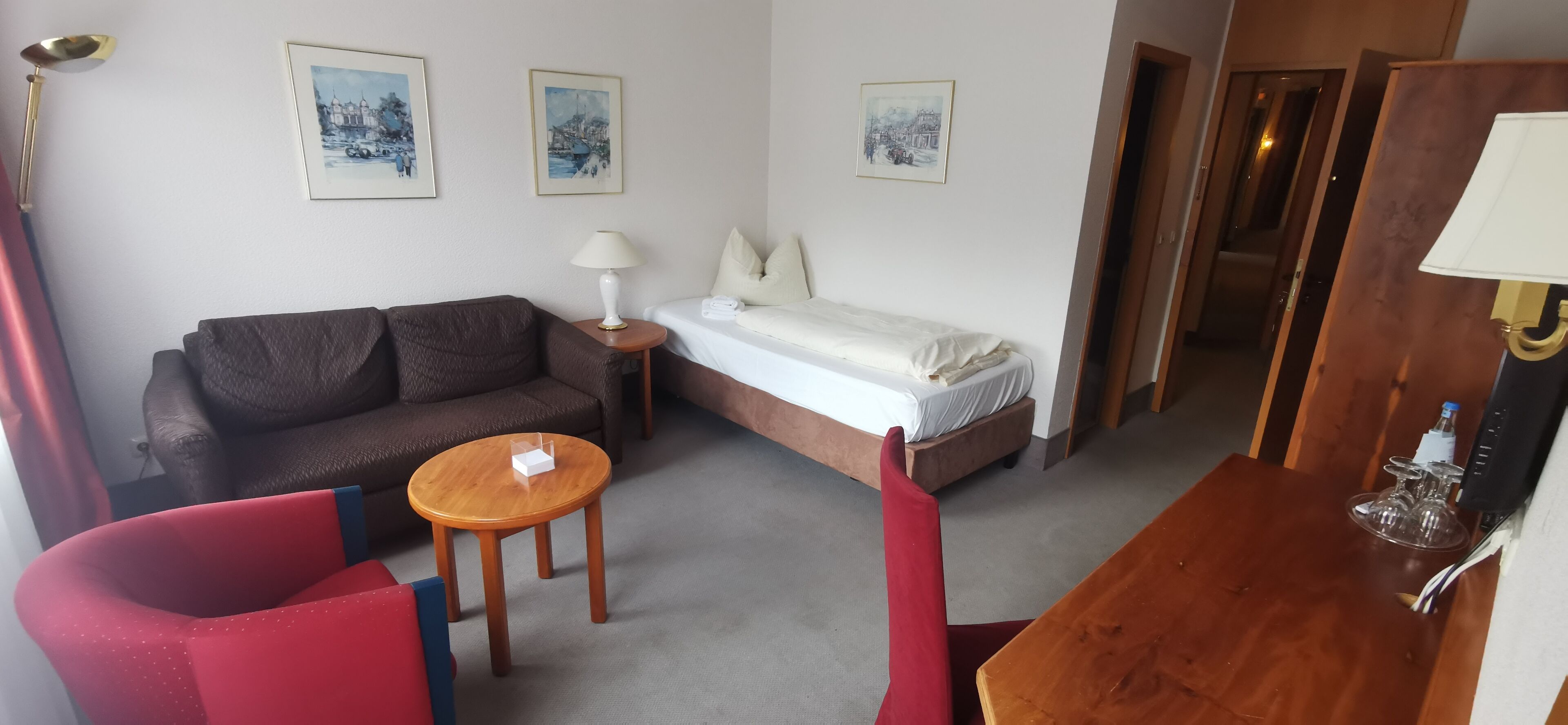 Une chambre d’hôtel compacte comprenant un lit simple, un canapé, une petite table et une chaise. Trois tableaux encadrés ornent le mur, et une lampe se trouve sur la table de chevet.