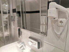 A modern bathroom with a glass shower enclosure, a hairdryer on a wall-mounted outlet, and a vanity with a mirror. - Heidehotel Soltauer Hof (Soltau)