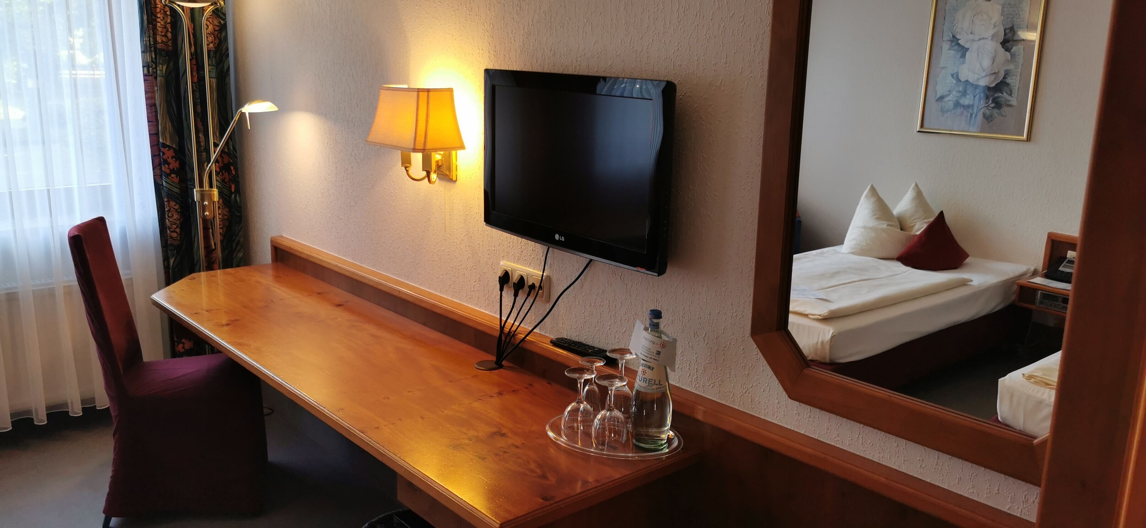 A hotel room with a wooden desk, a television mounted on the wall, a bed with white bedding, a lamp, and a bottle of water.