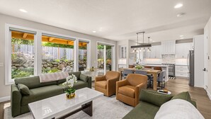 A modern living room with a sofa, armchairs, and a coffee table. There is a kitchen area with white cabinets and a dining table.