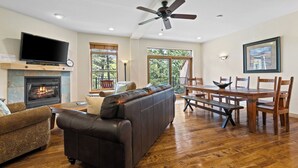 A spacious living room with a fireplace, a large flat-screen TV, a leather sofa, a dining table with chairs, and a ceiling fan.