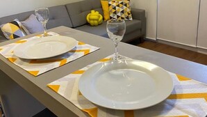 Een eethoek met een lange tafel gedekt voor twee personen, twee witte borden en twee wijnglazen. De tafel is afgedekt met een tafelkleed met een geel en wit patroon.