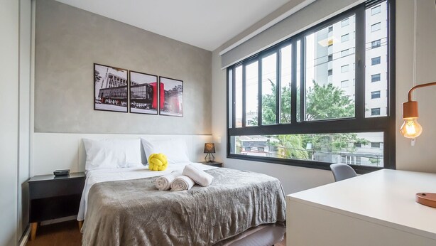Een moderne hotelkamer met een groot raam, een bed met een grijs gebreid plaid, een nachtkastje met een lamp en een bureau met een lamp.