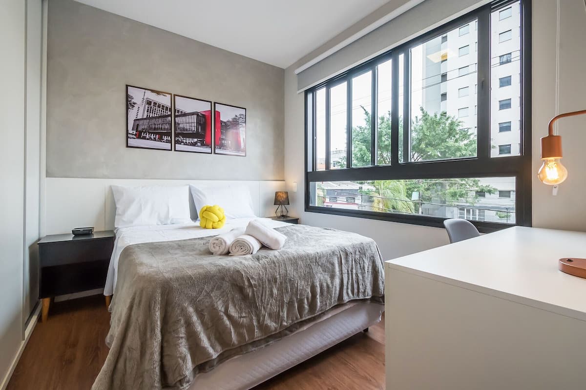 Une chambre d’hôtel moderne dotée d’une grande fenêtre, d’un lit recouvert d’une couverture grise à texture, d’une table de chevet avec une lampe et d’un bureau également équipé d’une lampe.