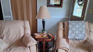 Un salon avec deux fauteuils beiges, une petite table ronde avec une lampe, un tableau encadré au mur et une porte décorée d’une guirlande.