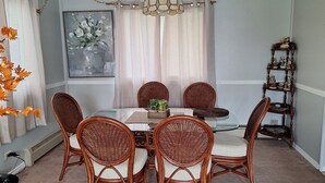 Un coin repas avec une table ronde et six chaises en osier, un lustre, un tableau au mur et une étagère en bois ornée de statuettes.