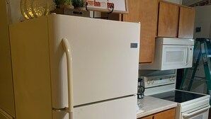 Une cuisine avec un réfrigérateur blanc, des armoires en bois, un four à micro-ondes et un four traditionnel. On peut voir un panneau sur le mur et une échelle à l’arrière-plan.