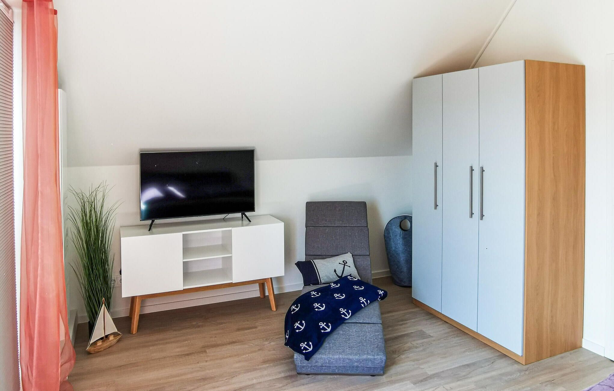 Un salon moderne avec un téléviseur à écran plat posé sur un meuble blanc, un canapé gris agrémenté d’un plaid bleu marine et une grande armoire.