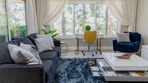 En moderne stue med en mørkegrå sofa, en blå lænestol, et hvidt sofabord og et stort vindue med gardiner.