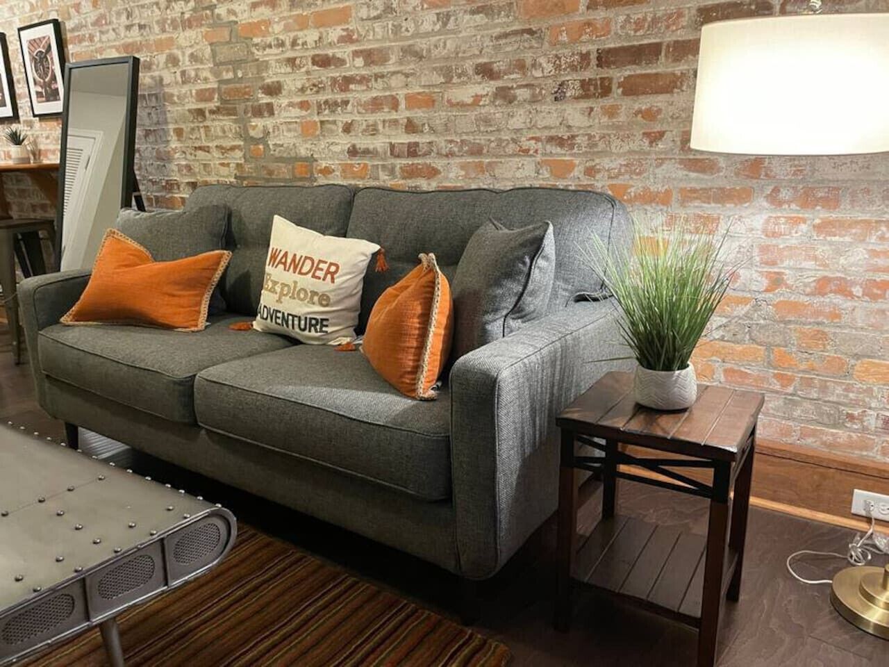 A grey sofa with orange and white cushions, a small wooden table with a plant, and a brick wall in the background.
