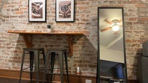 A room with a brick wall, a wooden shelf with two framed pictures, two metal stools, a tall mirror, and a ceiling fan. - Heart of Downtown 1 BR/1 BA - Hidden Gem (Norfolk)