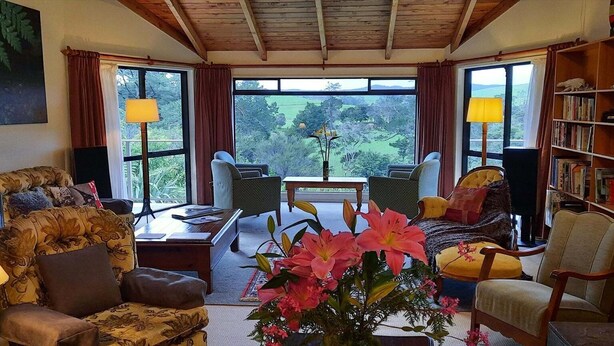 A cozy living room with a sofa, armchairs, a coffee table, and a bookshelf. Large windows offer a view of greenery outside. - Balcony river views and great value (Haruru)