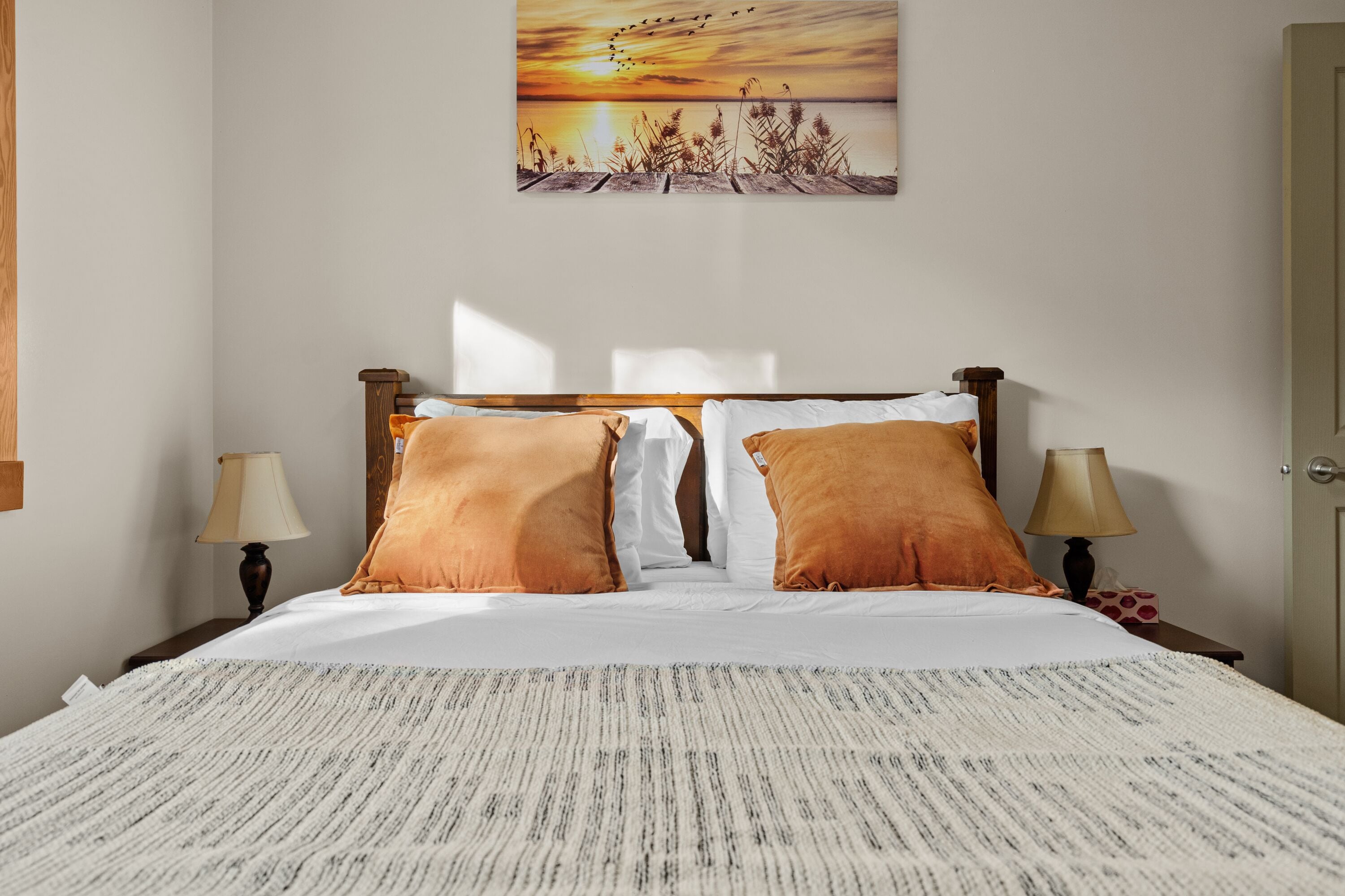 Een net opgemaakt bed met een bedsprei met patroon, twee bedlampjes en een schilderij van een zonsondergang aan de muur.