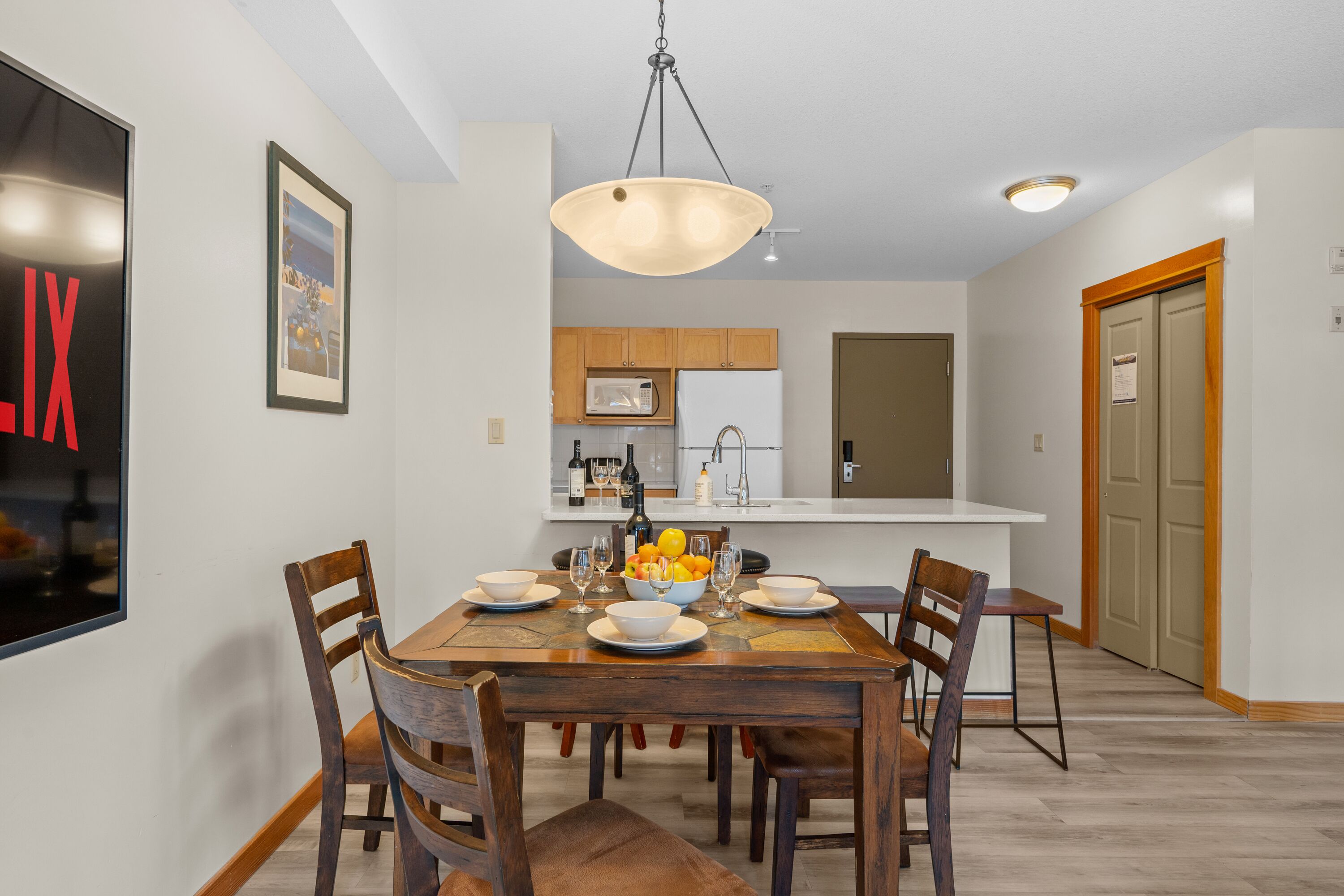Un comedor con una mesa de madera dispuesta para cuatro personas, rodeada de sillas, una lámpara colgante y una cocina al fondo.
