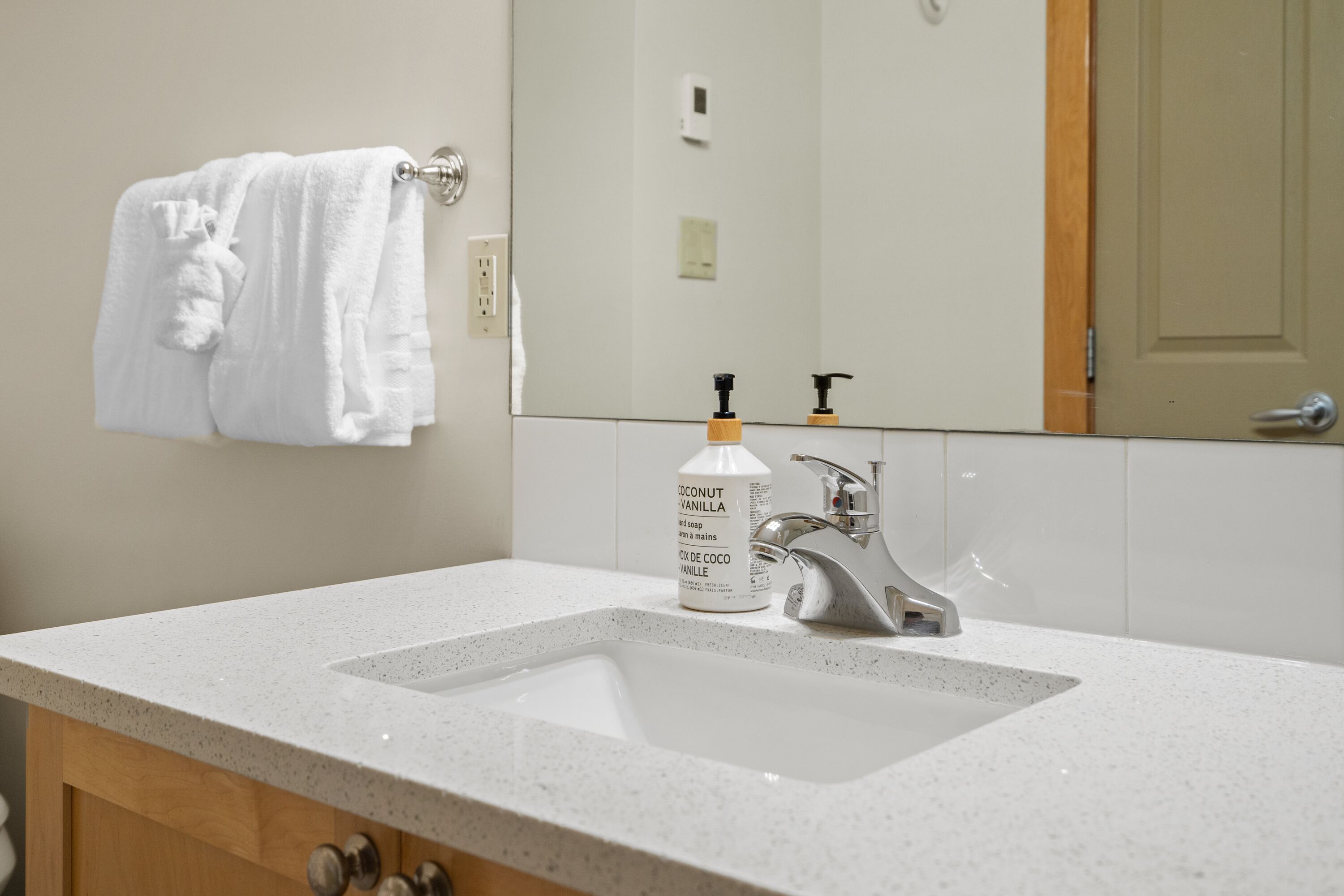 Une salle de bain avec un lavabo blanc, un miroir, une serviette suspendue à un porte-serviettes et un flacon de savon liquide pour les mains au parfum de vanille et de noix de coco.