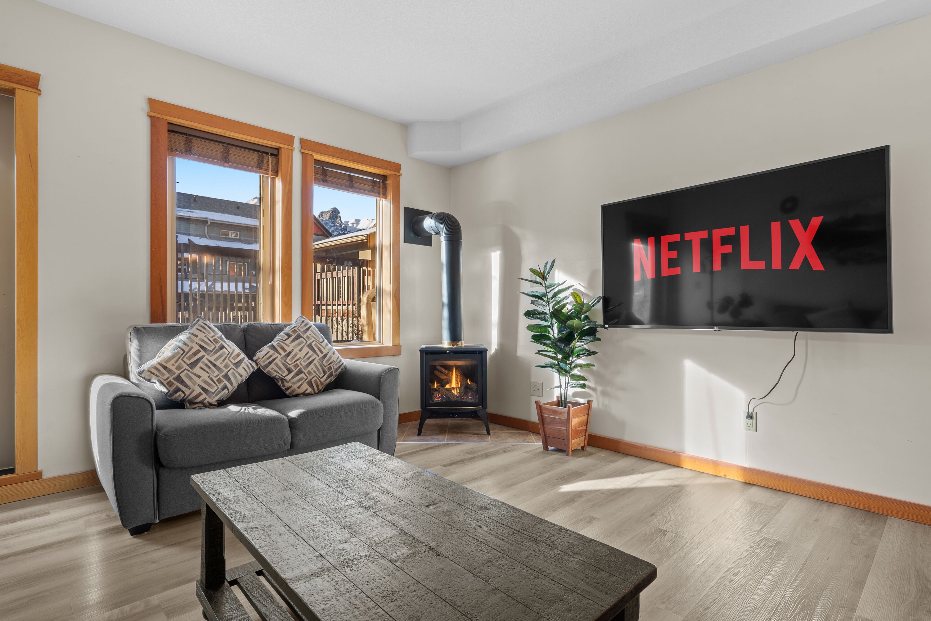 A living room with a gray sofa, a wooden coffee table, a fireplace, a potted plant, and a wall-mounted TV displaying the Netflix logo.