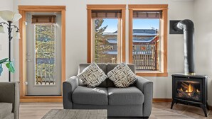 A living room with a gray sofa, two patterned pillows, a wooden floor, a fireplace, and a view through a window. - Heated outdoor Pool & Tubs 2 Bedroom & 2 Full baths 5 beds 9 Guests (Canmore)