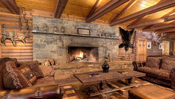 Interior de una cabaña de troncos con chimenea de piedra, muebles de cuero y adornos de asta.