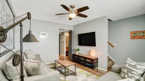 Una sala de estar moderna con un sofá, una mesa de centro y un televisor montado en la pared. Se observa un ventilador de techo, una lámpara y una estantería.
