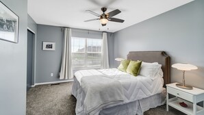Un dormitorio con cama, ventilador de techo, mesita de noche y ventana con cortinas.