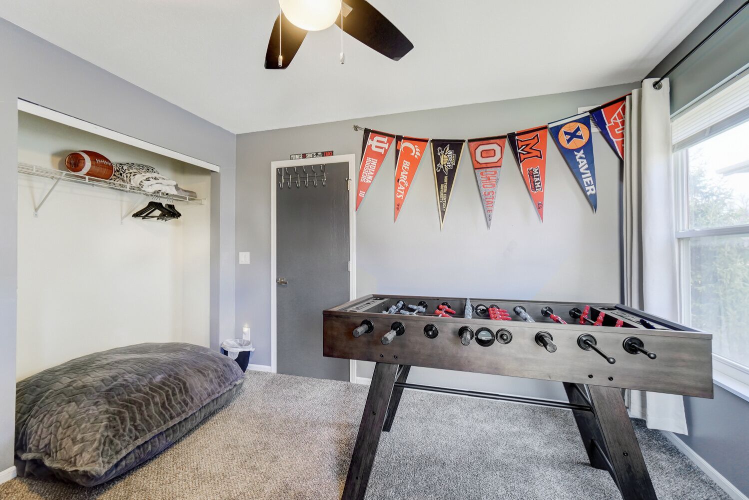 A room with a foosball table, a gray carpet, a gray wall, a window, and a ceiling fan.
