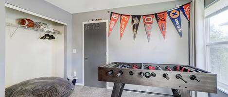 A room with a foosball table, a gray carpet, a gray wall, a window, and a ceiling fan.