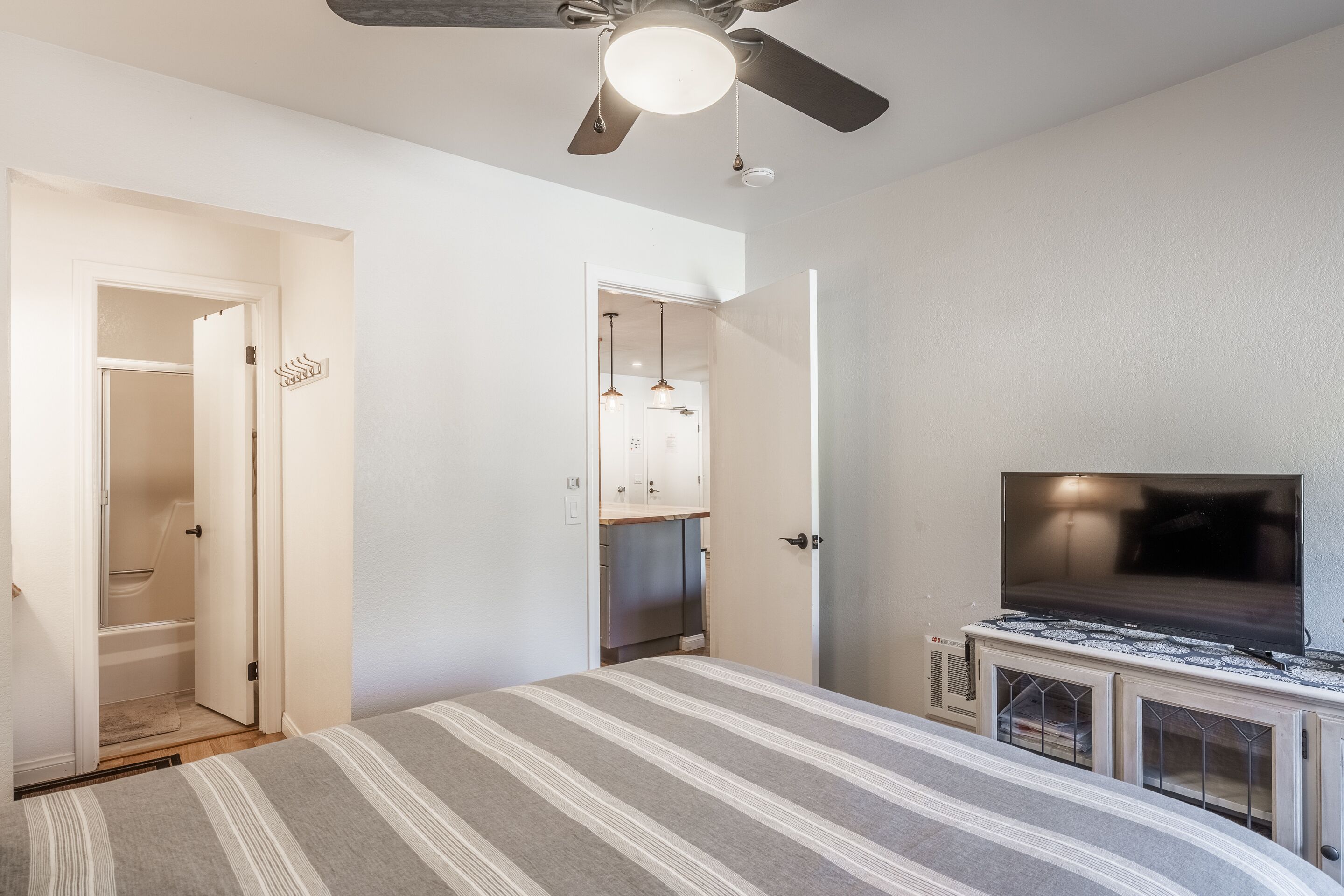 A bedroom with a bed, a ceiling fan, a TV, and a kitchenette.