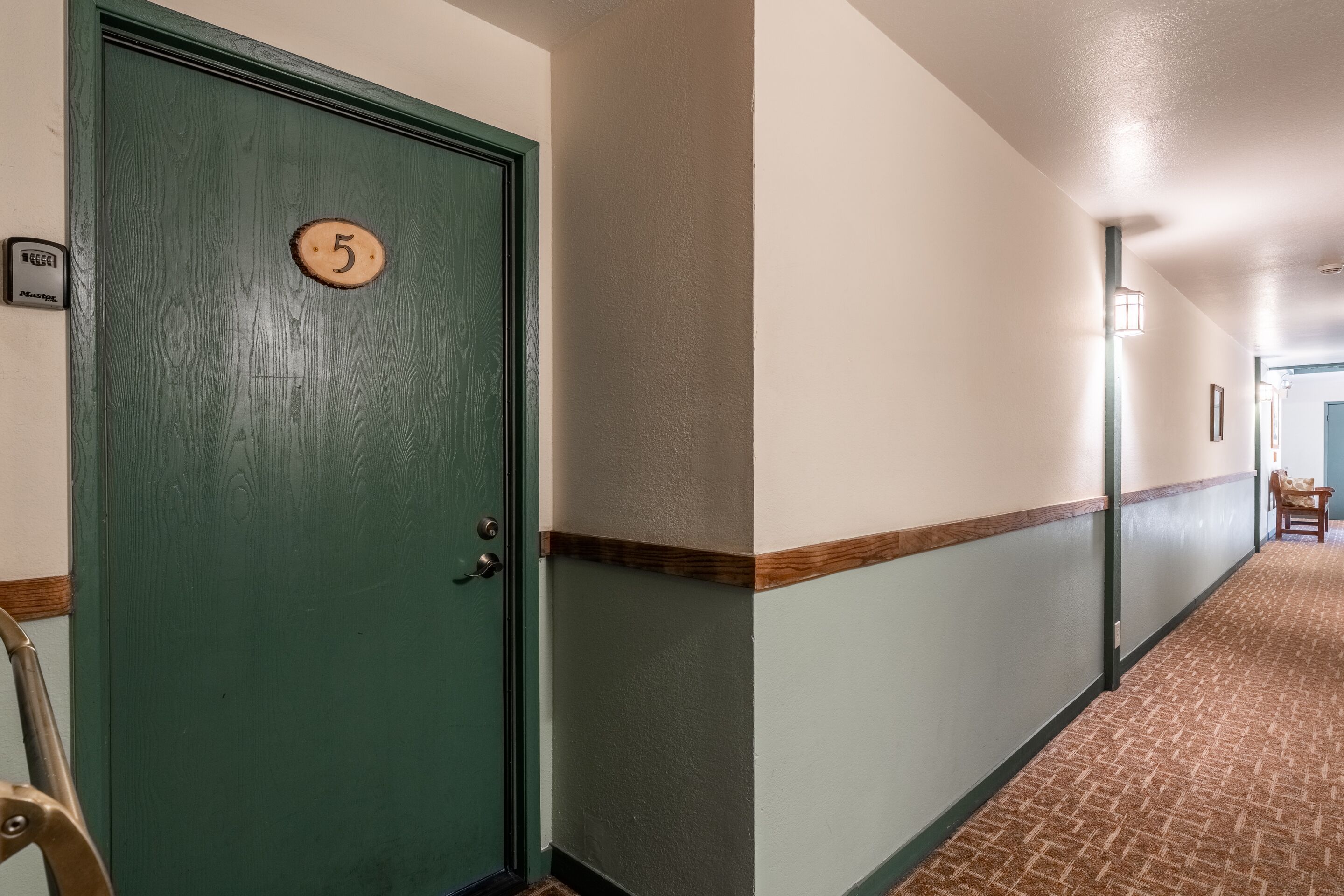 Un couloir avec une porte verte portant le numéro 5, un interrupteur et une chaise.