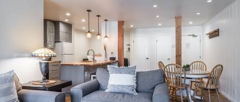 Ein modernes Wohnzimmer mit grauem Sofa, einem Esstisch mit Holzstühlen und einer Küchenzeile mit Schränken und einem Kühlschrank.