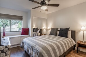Un dormitorio con una cama grande, ventilador de techo, lámparas de noche, una ventana con persianas y vistas al exterior.
