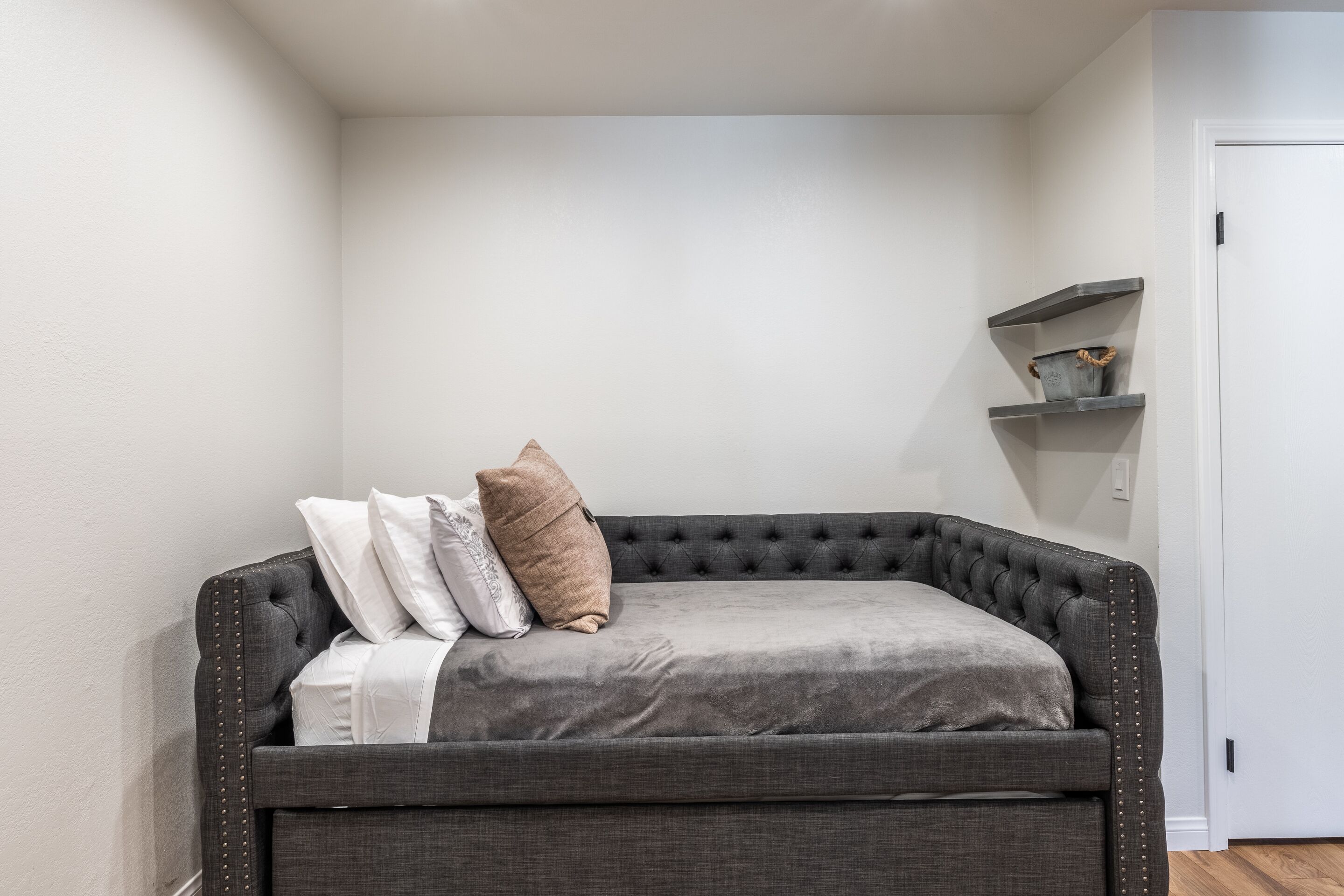 Et hjørneværelse med en grå polstret sofa, en hvid dør og en lille reol med dekorative genstande.