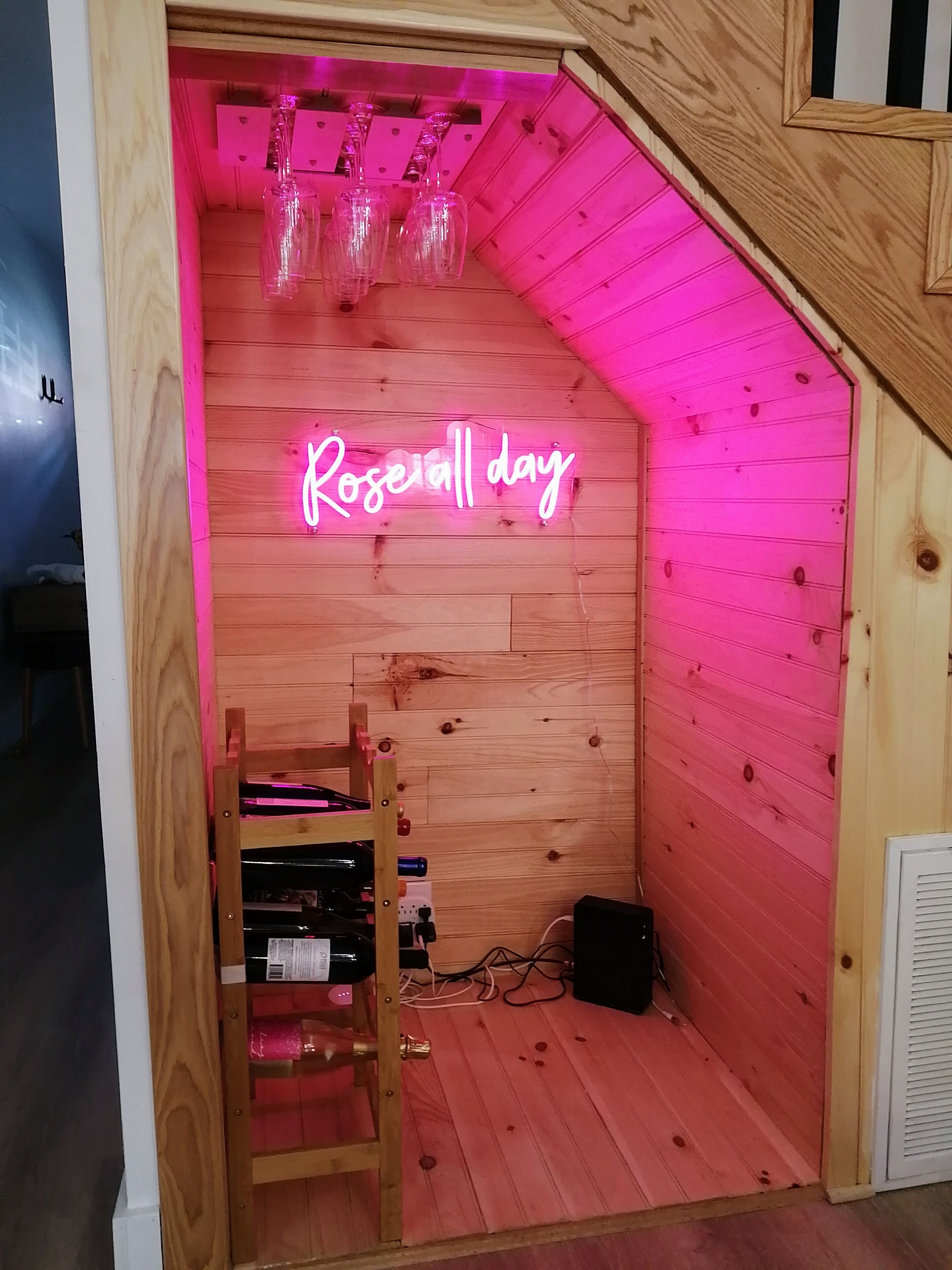 Une pièce chaleureuse aux murs en bois et au plafond en pente, agrémentée d’un néon indiquant « Roseal day » et d’une petite étagère où sont posées des bouteilles de vin.