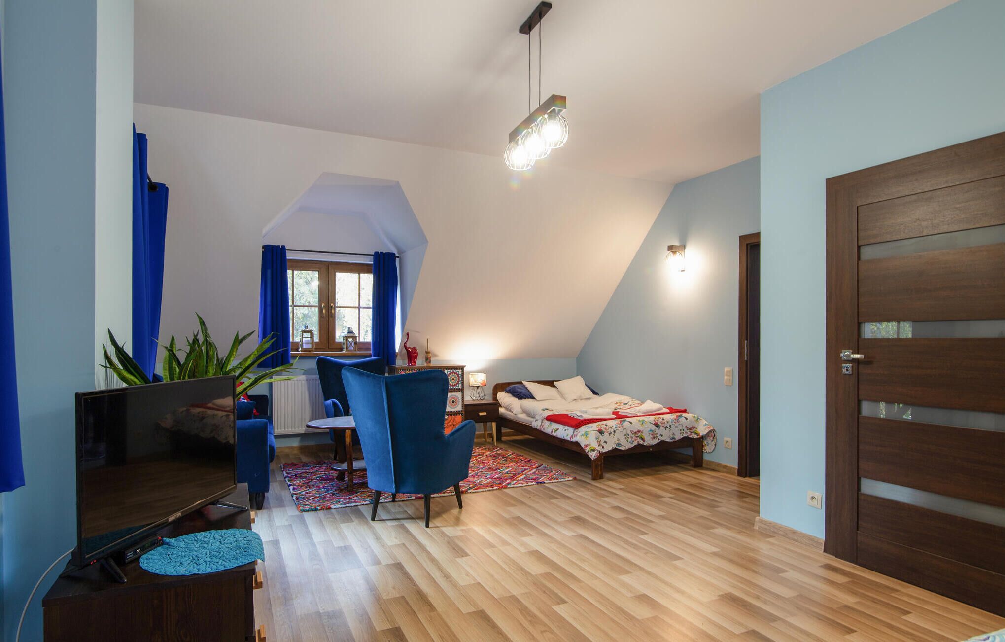 A bedroom with a sloped ceiling, a bed with a floral bedspread, a desk with a chair, a television, and a door with a frosted glass panel.
