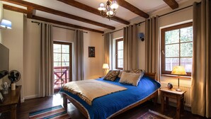 Un dormitorio con una cama grande, vigas de madera, una araña y dos ventanas con cortinas.