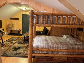 Una cama litera con colcha escocesa, una mesa de comedor de madera con sillas y un banco en una habitación con paredes y techo inclinados.