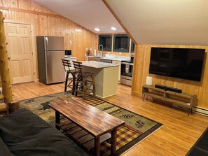 A compact kitchen with a refrigerator, stove, and sink, a dining area with a wooden table and chairs, and a television on the wall.