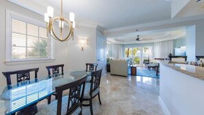 Amplia sala de estar con mesa de vidrio, sillas de madera oscura, sofá color beige y una zona de cocina con encimera y electrodomésticos.