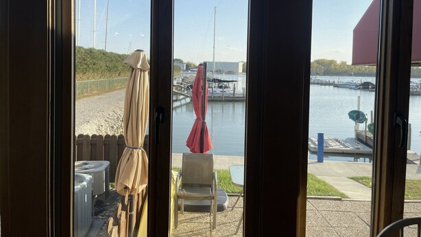Vue depuis l’intérieur d’une pièce, donnant sur une marina où des bateaux sont amarrés, avec une clôture en bois, une chaise et un parasol à l’extérieur.