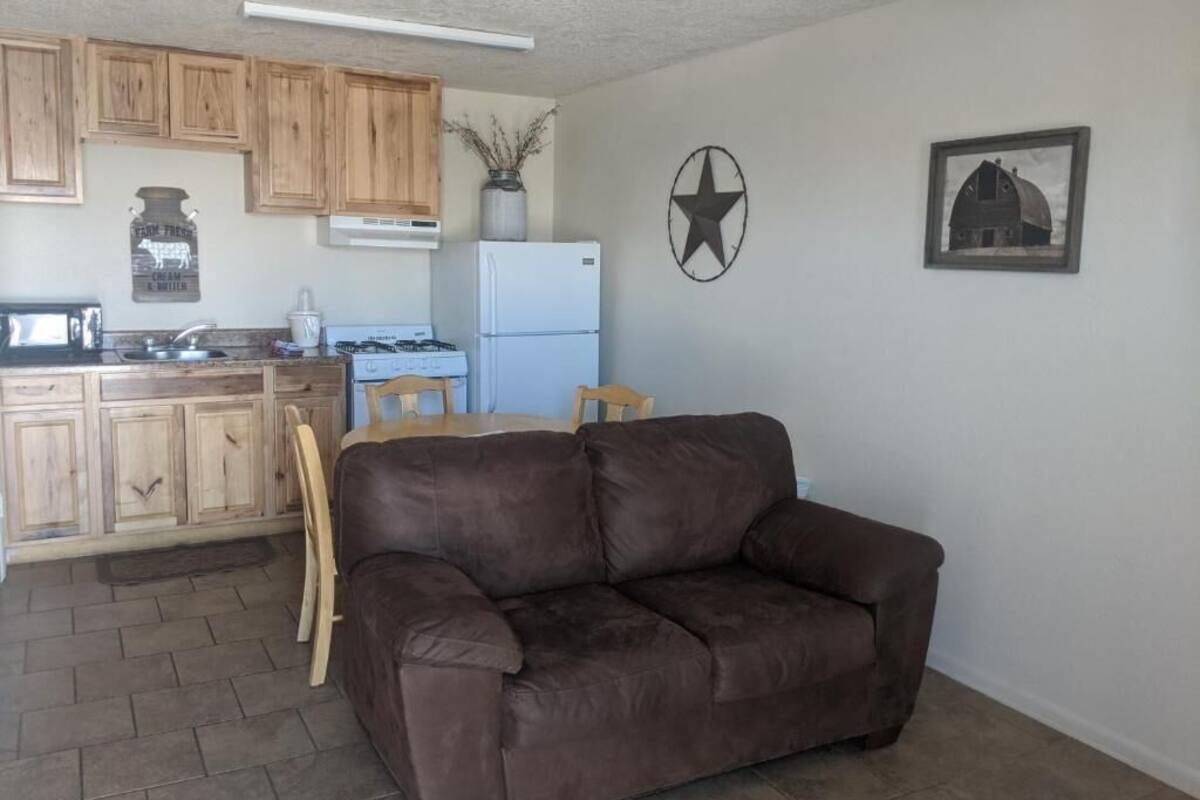 A living room with a brown leather sofa, wooden cabinets, a kitchen area with a refrigerator, stove, and sink, and a wall with a star and barn picture.