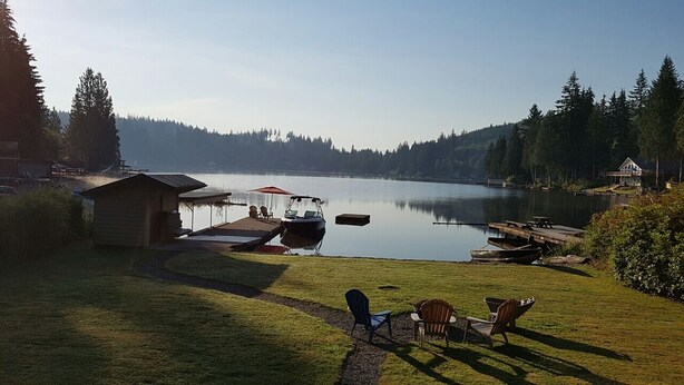 Una propiedad junto al lago con muelle, una cabaña pequeña y un área de césped con mobiliario exterior.