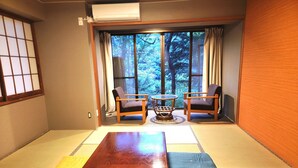 Una habitación japonesa tradicional con suelo de tatami, una mesa de madera baja, dos sillas y vistas a un bosque a través de amplios ventanales.
