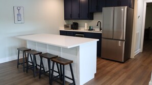 Eine moderne Küche mit dunklen Schränken, einem Edelstahlkühlschrank, einer weißen Kochinsel mit Barhocker und einem Toaster.