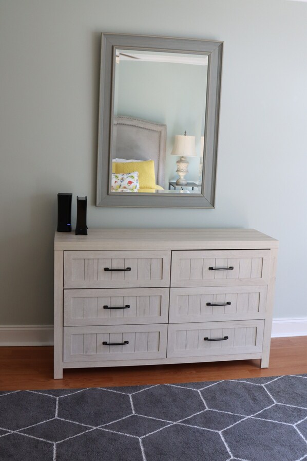 A light-colored dresser with four drawers, a mirror above it, and a hexagonal-patterned rug on the floor.