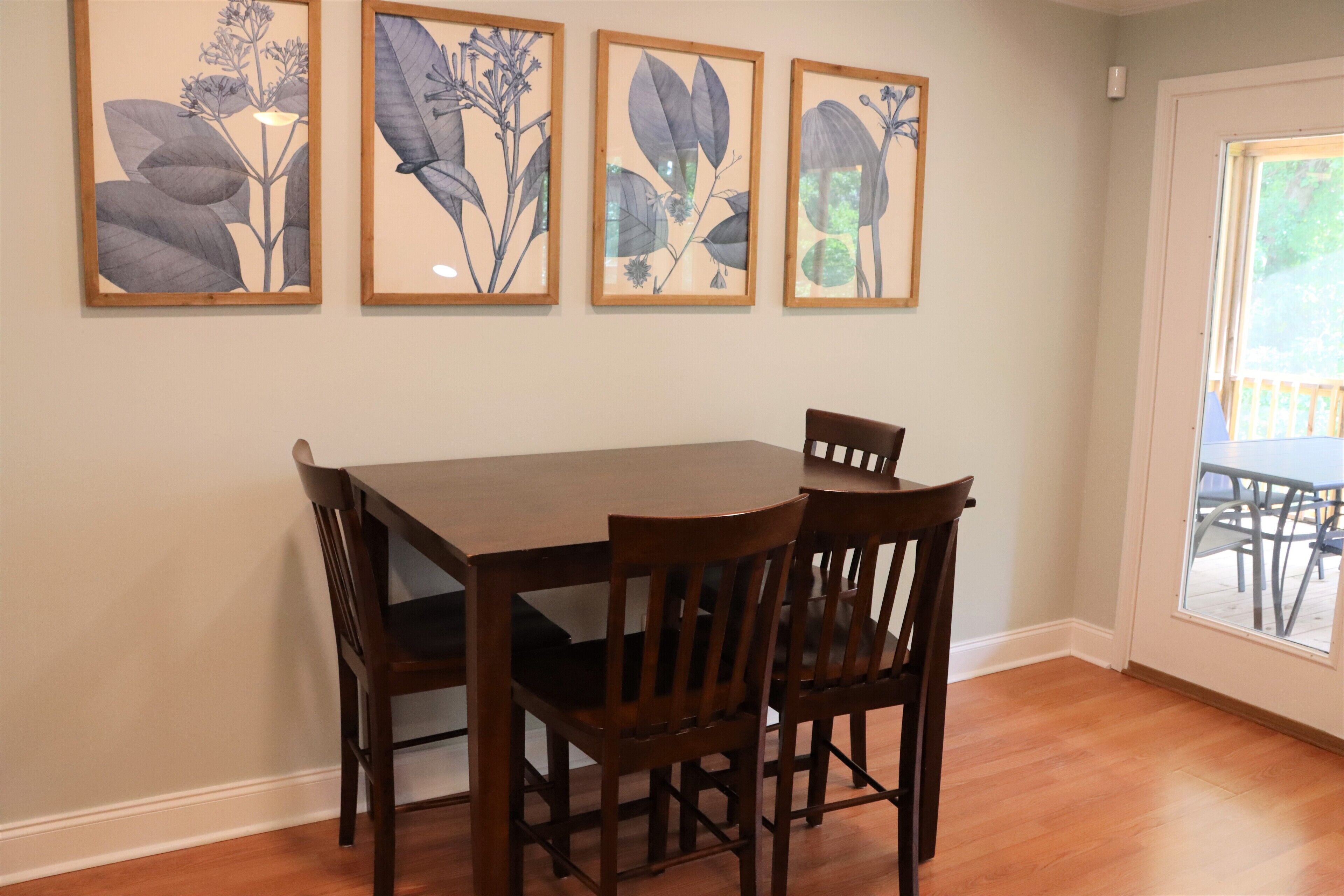 A dining area with a wooden table and chairs, framed botanical prints on the wall, and a view through a glass door.